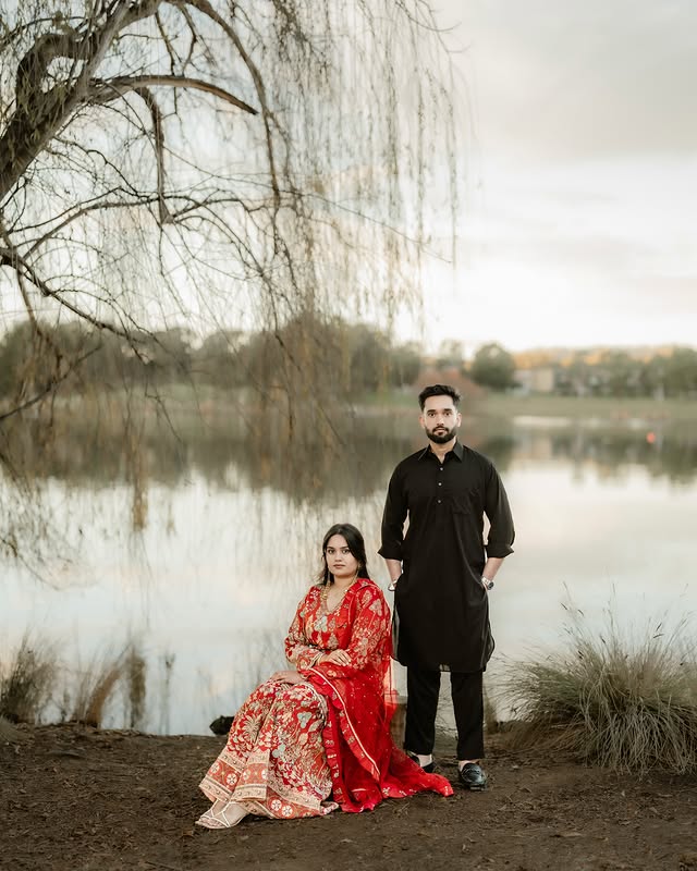 Bride in richly embroidered red attire with her partner by a lake
